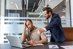 A frustrated manager confronts a stressed employee in an office setting, pointing at a laptop. The scene depicts a work-related conflict between colleagues. photo