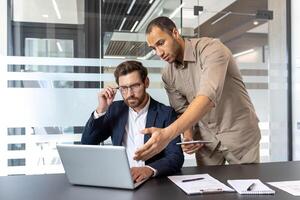 Two businessmen are collaborating in a modern office, discussing a project displayed on a laptop. One points to the screen. photo