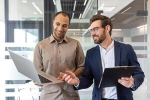 Two businessmen smile while working on a laptop in a modern office setting, discussing. One holds a clipboard. Collaboration is shown. photo