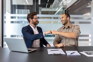Two men, likely colleagues, bump fists in a modern office environment. A laptop and documents are on the table, indicating a business meeting or discussion. photo