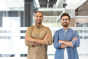 Two confident men stand in an office setting, arms crossed, smiling at the camera. They showcase a professional demeanor. photo