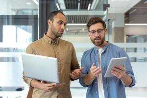 Two professional men collaborate in a modern office, using a laptop and tablet for discussion. They appear focused and engaged in their work. photo
