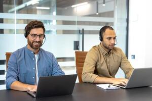 Two customer service representatives wearing headsets are working at desks using their laptops in a modern office setting. photo