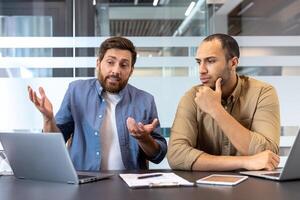Two diverse business professionals discuss something in a modern office environment, using a laptop and other devices. photo