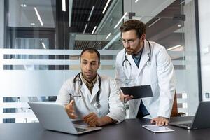 Two doctors in white coats are discussing something on a laptop, a clipboard, and a tablet in a modern office setting. photo