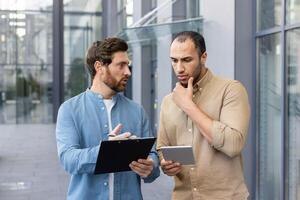 Two men are engaged in a discussion outdoors. One holds a clipboard, while the other considers a tablet in their hands. photo