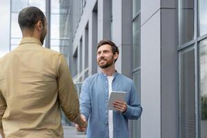 Two business partners shake hands in front of a modern office building, smiling. One holds a tablet. photo