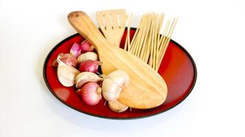 wooden spatula, skewers, and shallots and garlic in a red plastic plate on a white background photo
