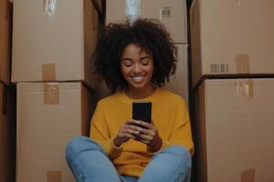 Smiling young woman sits among cardboard boxes while using her mobile phone during the moving process to her new home photo