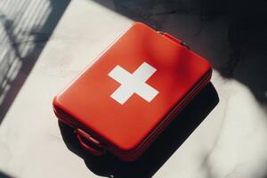 Red first aid kit with white cross placed on a surface, showing shadows and details in a bright natural light during the day photo