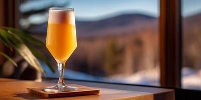 A golden cocktail in a tall glass, topped with foam, sits on a wooden surface by a window with a mountain view photo
