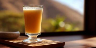 A tall, stemmed glass filled with a light orange smoothie, set on a wooden tray, with a blurred view of a landscape outside a window photo