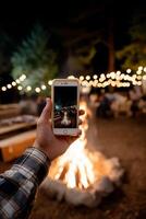 A person holding up a smartphone in front of a campfire photo