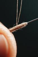 Close up view of thread being pulled through eye of needle, detail of beige yarn and metal with dark background. photo