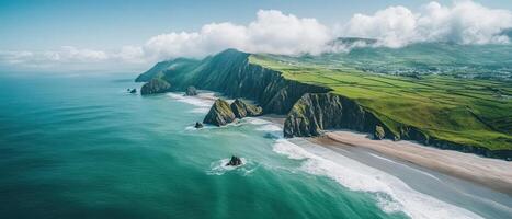 Coastal cliffs meet the sea. Green fields above cliffs. Waves reach sandy shore. Mist and clouds hug mountain peaks. Azure ocean water. photo