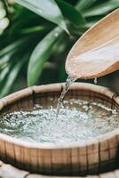 Clear water flows from bamboo spout into round wooden basin in lush green natural setting forming droplets and splashes of liquid photo