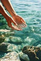 Hands touch clear water. Ripples spread over surface. Smooth stones visible beneath the surface. Sunlight reflects on rocks in shallow water. photo
