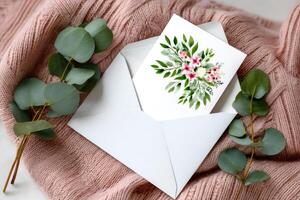 Greeting card with floral design emerging from white envelope lying on pink knitted sweater with eucalyptus leaves photo