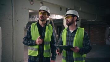dois caucasiano capatazes rapazes construtores ao controle industrial trabalhos dentro construção técnicos homens arquitetos construção cooperação faz-tudo dentro segurança uniforme discutindo engenheiros conversa walkie talkie planejamento video