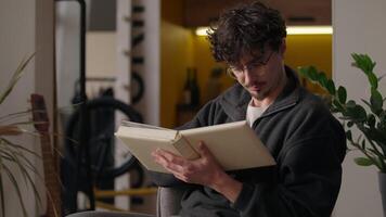 Focused Caucasian guy reading book at home hipster male read story literature paper knowledge reader in glasses studying information library hobby man learn homeowner holding textbook notebook leisure video