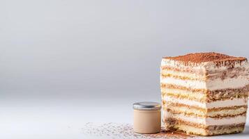 A delicious slice of layered tiramisu cake sits on a white surface next to a small jar of cream dusted with cocoa powder against a light gray background. photo