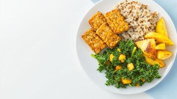 A healthy and delicious vegan meal featu tempeh, roasted potatoes, kale, quinoa, and chickpeas is served on a white plate against a light blue background. photo