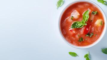 A refreshing bowl of chilled gazpacho soup garnished with fresh basil and diced vegetables sits on a light blue background offe a vibrant summer food image perfect for culinary photo