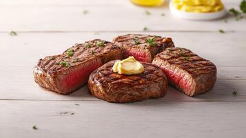 Four delicious grilled filet mignon steaks are artfully arranged on a rustic white wood table with herb garnish and a pat of butter melting on top. photo