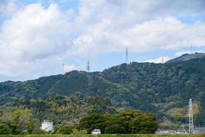 A scenic landscape featuring lush green mountains under a partly cloudy sky. Tall electricity transmission towers line the ridges photo
