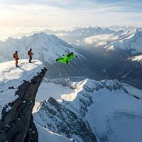 A wingsuit flyer jumps from a snowy mountain peak while two figures watch, with a vast mountain range in the background photo