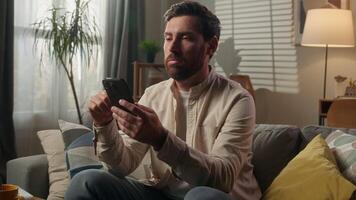 Sad upset frustrated man Caucasian businessman guy bored using mobile phone on couch sofa worried disappointed male homeowner reading news cellphone browsing message scam scrolling smartphone at home video