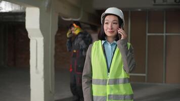 multiétnico construtores coreano mulher caucasiano homem trabalhando construção Civil engenheiros dentro capacete de segurança uniforme capataz inspetor falando Móvel telefone ligar comunicando o negócio projeto construção desenvolvimento local video