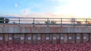 Reinforced concrete retaining wall with pile numbers beside a reservoir at sunset. Infrastructure structure designed to prevent soil erosion and flooding, bridge over the river video