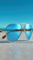 Stylish sunglasses resting on a sandy beach with clear blue skies and the ocean in the background during midday photo