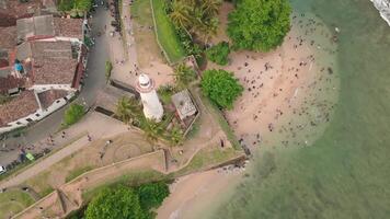 Aerial top down view people on the beach swim relax by Galle fort Lighthouse in Sri Lanka, showing coastal heritage and islamic lifestyle, white architecture, islamic colonial architecture buildings video