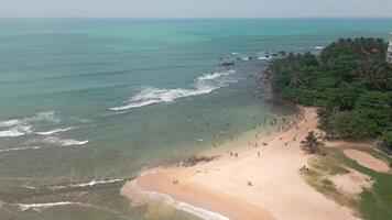Aerial panorama view crowded beach in Tangalle, Sri Lanka, where tourists enjoy swimming and sunbathing along the tropical coastline. A lively vacation scene with turquoise waves and palm-lined shore video