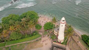 Drone fly over view of Sri Lankans enjoying a swim in the ocean near the iconic white lighthouse at Galle Fort. The scene blends historic architecture with daily coastal life and island leisure video