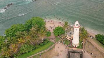 Aerial circular view of Sri Lankans enjoying a swim in the ocean near the iconic white lighthouse at Galle Fort. The scene blends historic architecture with daily coastal life and island leisure video