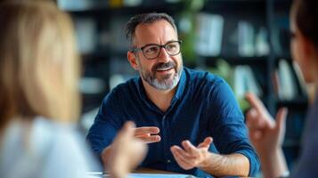 A man with glasses and a beard is sitting at a table, smiling and gesturing with his hands while talking to someone. photo