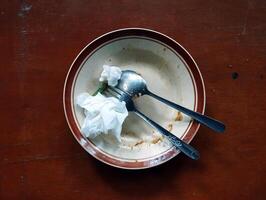 An empty plate with leftover food, a spoon, a fork, and some used tissues placed on a brown table. photo