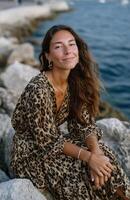 Beautiful woman in leopard-print dress relaxing on rocks by the sea during golden hour photo