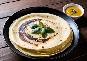 Stack of Rava dosa with basil garnish on a black plate next to a bowl of yellow curry on a wooden table photo