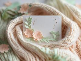 Blank Card with Peach Paper Flowers and Green Leaves on Cozy Knitted Scarf Background. photo