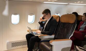 Cheerful businessman in black suit using laptop computer while talking on cellphone in airplane cabin photo