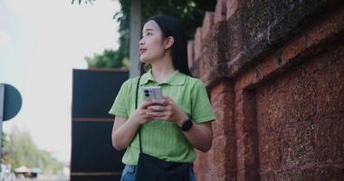 Handheld low angle view shot, Young Asian woman in green shirt and blue jeans looks at GPS on mobile app and looks at surrounding view while walking on sidewalk beside brick wall. video