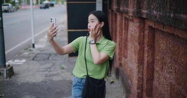 Handheld high angle view shot, Cheerful young woman happily takes a selfie while walking along a sidewalk next to a brick wall. video