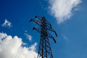 Electrical transmission tower silhouette against bright blue sky with scattered white clouds, high voltage power line pylon photo