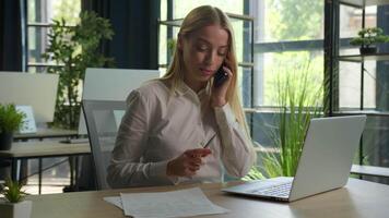 Smiling girl Caucasian business woman multitasking working in office typing on laptop talking phone writing papers documents consulting client support customer mobile call multitask businesswoman work video