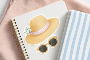 Stylish straw hat with pink ribbon and floral accent placed on a notebook beside round sunglasses, creating a chic summer aesthetic for design inspiration and lifestyle imagery photo