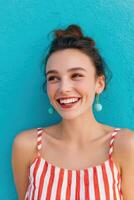 Female software developer with a joyful smile, wearing a striped top, stands against a vibrant turquoise wall, showcasing a cheerful and confident demeanor in a modern workspace photo